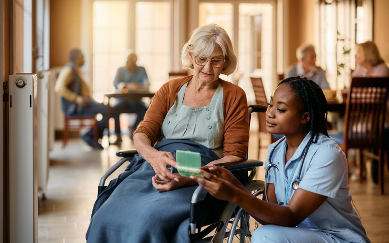 Young black nurse giving pills to senior woman in wheelchair at nursing home.